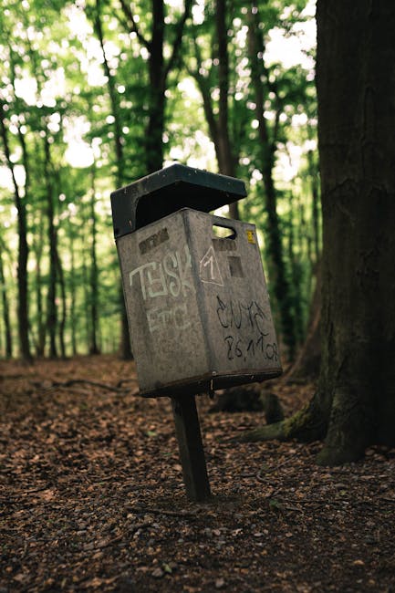 Two green plastic rubbish bins with hinged lids are positioned side by side on a city sidewalk, close to the edge of a paved area with some loose debris and dried leaves around their base. The bins ar